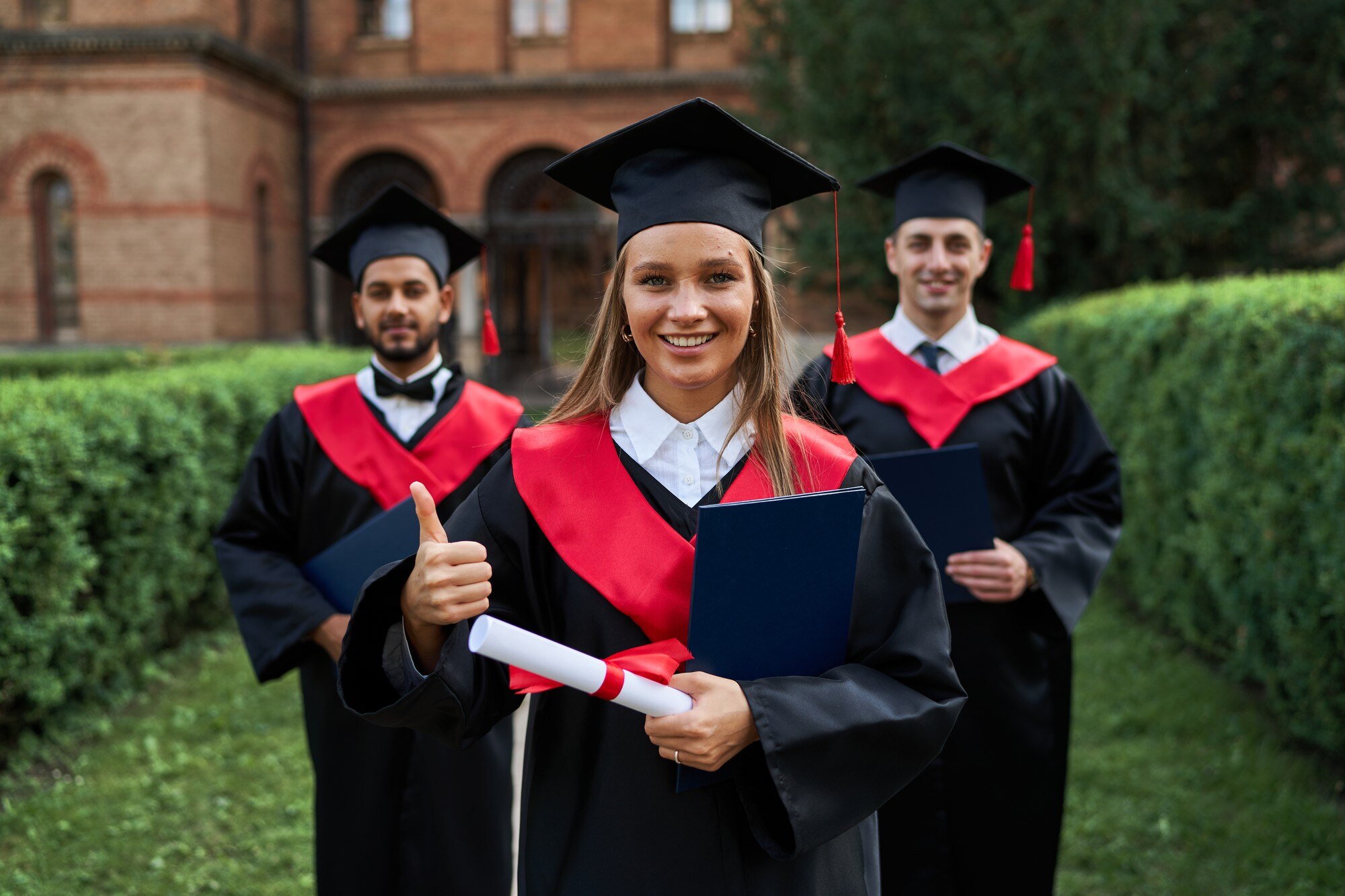 graduate-female-shows-like-with-her-friends-graduation-gowns-holding-diploma-smiling-camera_496169-1308